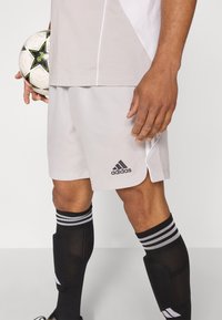 Vaaleanharmaat Adidas-shortsit, joissa on musta logo ja sivuaukot. Mustat polven yli ulottuvat sukat valkoisilla raidoilla. Henkilö pitää jalkapalloa.