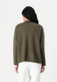 Olijfgroene, lange mouw sweater met een losse pasvorm, geribbelde halslijn en een iets langere achterkant. Geselecteerd met witte broek.