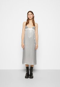 Vestido de slip de cetim prateado com decote em V e alças finas, caindo até à metade da panturrilha. Combinado com botas de tornozelo de sola grossa pretas.