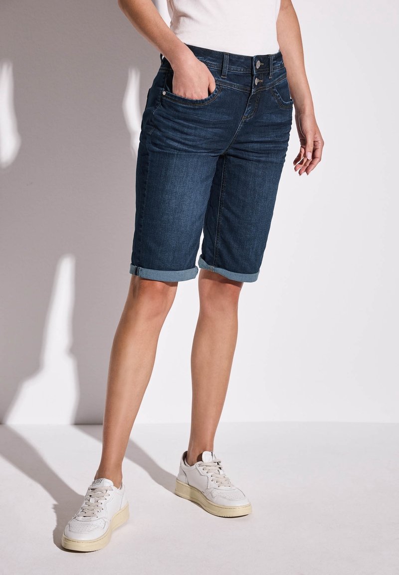 Mörkblå denimshorts med uppvikta fållar, sidofickor och dragkedja. Kombineras med vita sneakers med en texturerad design.