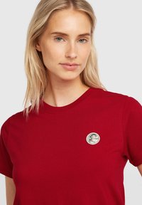 Red short-sleeve t-shirt made of cotton, featuring a round, embroidered logo patch in white and black on the chest. Smooth texture.
