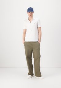 Witte poloshirt met een klein logo, olijfgroene wijde broek, witte sneakers en een blauwe pet, staande tegen een effen achtergrond.