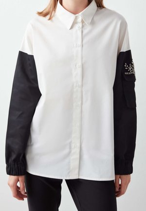 Femme portant une chemise blanche à boutons avec des manches noires munies de poignets élastiques et une poche ornée sur le bras gauche.