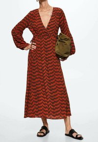 Frau in einem langärmligen Kleid mit rot-schwarzem Zickzackmuster, die eine olivgrüne, lässige Tasche hält und schwarze Sandalen trägt.