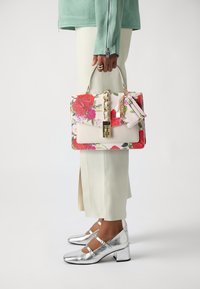 Sac à main floral avec des roses roses et rouges, matériel en chaîne dorée et une étiquette détachable, associé à des escarpins Mary Jane argentés et un pantalon clair.