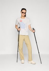 Ljust grå t-shirt med blå grafisk design och röd sol, i kombination med beige byxor och vita vandringsskor. Håller i vandringsstavar.