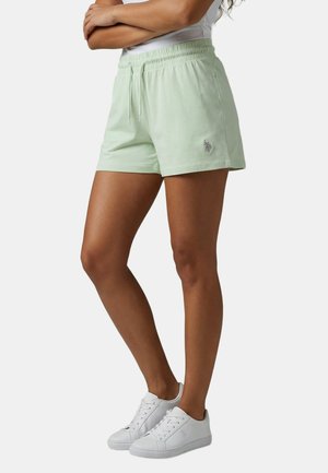 Frau trägt hellgrüne Shorts mit Kordelzug und weiße Sneakers, steht mit verschränkten Armen vor einem schlichten Hintergrund.