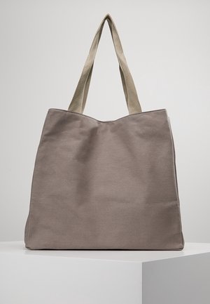 Sac fourre-tout en toile beige avec de larges poignées en tissu beige clair, debout sur une surface blanche contre un fond gris clair uni.