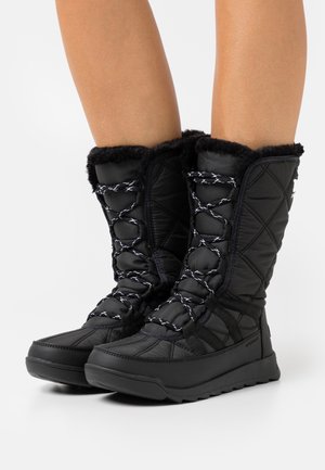 Bottes d'hiver noires matelassées avec doublure en fourrure et lacets réfléchissants portées sur des jambes nues, présentées sur un fond blanc.