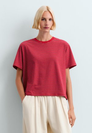 Femme blonde aux cheveux courts portant un t-shirt à manches courtes rayé rouge et blanc et un pantalon ample beige clair, debout devant un fond uni.