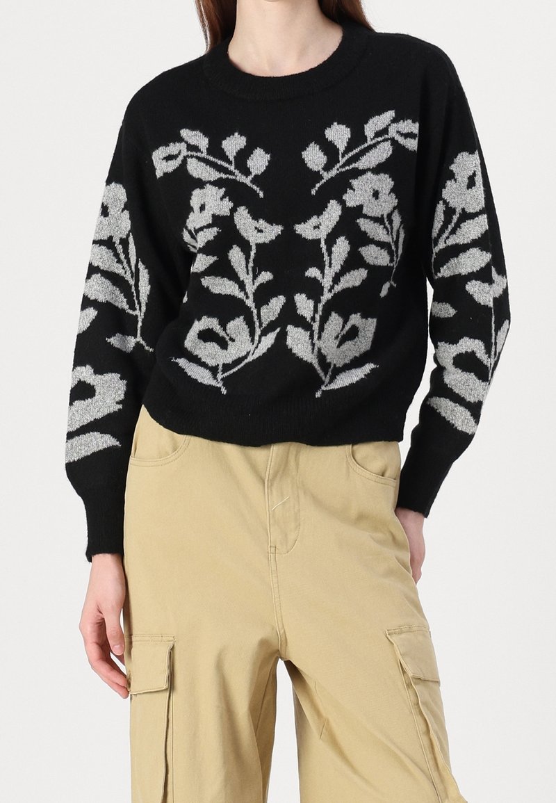 Pull noir avec motifs floraux gris clair sur le devant et les manches, porté avec un pantalon cargo beige.