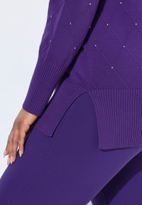 Ruka spočívající na fialových legínách, oblečená ve fialovém svetr s ribonovaným lemem a diamantovým vzorem zdobeným malými korálky.