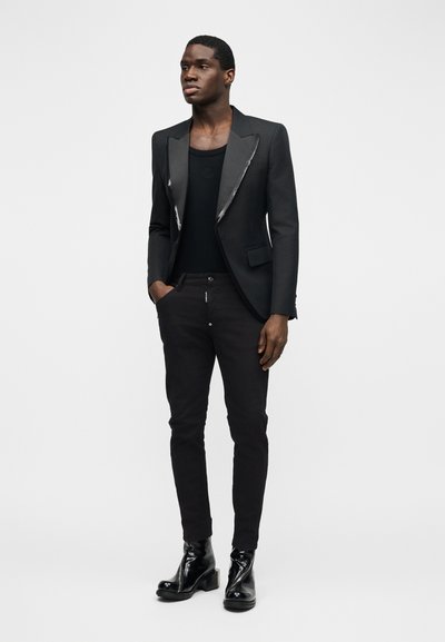 Νεαρός άνδρας που φοράει σακάκι με σατέν πέτο σε μαύρο χρώμα, μαύρο πουκάμισο, στενά μαύρα παντελόνια και γυαλιστερά μαύρα μποτάκια μέχρι τον αστράγαλο, στέκεται με το ένα χέρι στην τσέπη.
