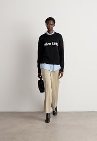 Person geht und trägt einen schwarzen Calvin Klein Pullover über einem hellblauen Hemd, beigen Hosen, schwarzen Stiefeln, klare Brille und hält eine kleine schwarze Tasche.