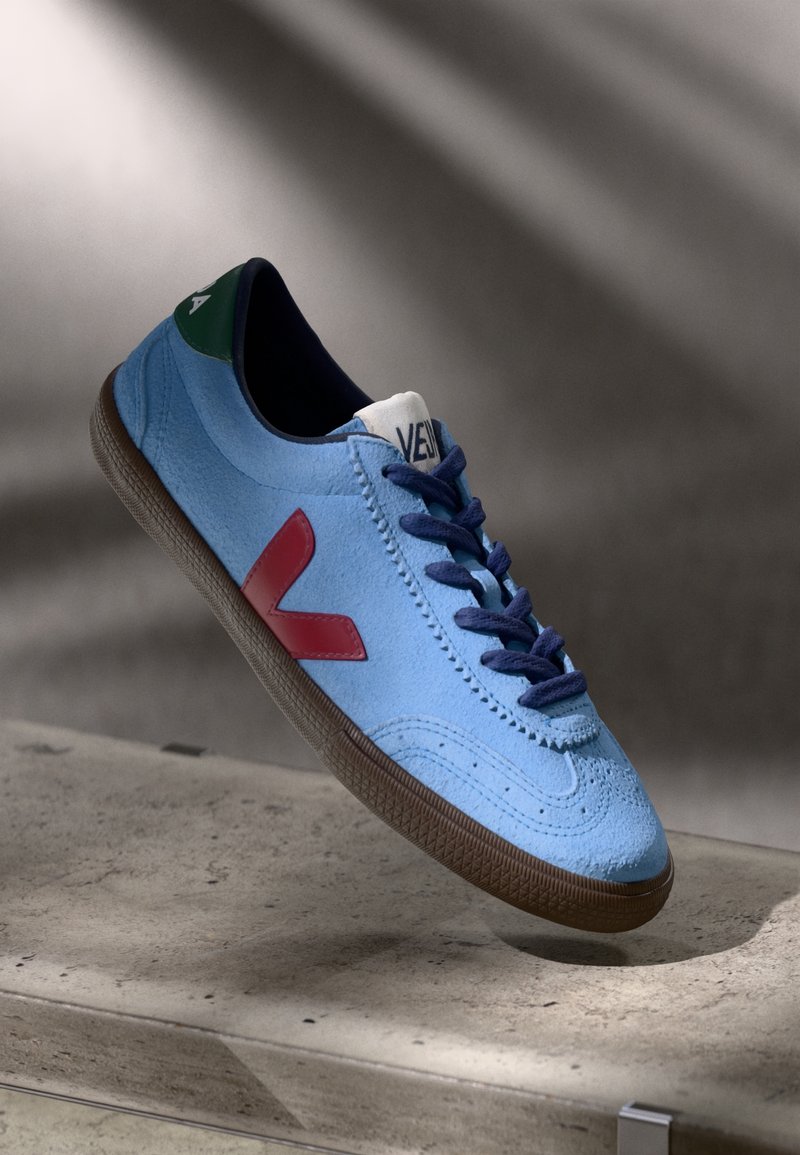 Baskets en daim bleu clair avec logo latéral rouge, lacets bleu foncé, semelle marron et détail vert au talon flottant au-dessus d'une surface en béton.
