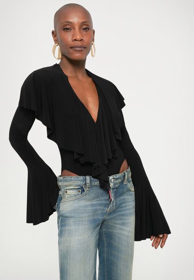 Γυναίκα με ξυρισμένο κεφάλι, φοράει μεγάλα κρίκους σκουλαρίκια, μαύρη μπλούζα με βαθύ V και φουσκωτά μανίκια, καθώς και ανοιχτόχρωμο τζιν, μπροστά σε λευκό φόντο.