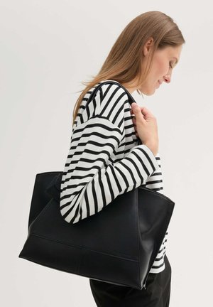 Mujer con cabello largo, vestida con camiseta de rayas blancas y negras, llevando un gran bolso tote de cuero negro sobre el hombro.