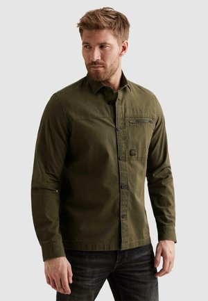 Moški, oblečen v olivno zeleno srajco z gumbi in žepom na prsih z zadrgo ter temne kavbojke, gleda levo proti enobarvnemu ozadju.