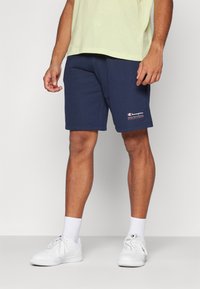 Navy modri bombažni šortki z elastičnim pasom, ki imajo na levem delu noge bel logotip "Champion" ter sproščen kroj. Bele superge in nogavice.