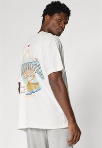 Camiseta de algodón blanca con un estampado gráfico colorido en la parte posterior que presenta un diseño de barco, acentos azules y amarillos, y un corte holgado.