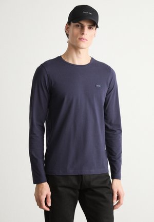Maglietta a maniche lunghe blu navy in morbido cotone con collo rotondo e piccolo logo ricamato sul petto. Abbinata a pantaloni neri e un berretto nero.
