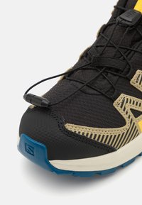 Svart og beige terrengsko med et teksturert mesh-overdel, elastisk snøresystem, gummikappe på tåen og en blå såle. Har stripete designelementer.
