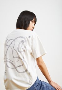 Hvid kortærmet t-shirt med et gråt abstrakt kæde-design på ryggen, med runde detaljer og en afslappet pasform.