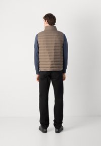Beige puffer vest met horizontale stiksels, hoge kraag en een gladde textuur. Draag het over een blauwe longsleeve, gecombineerd met zwarte broek.