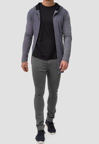 Grauer Langarm-Hoodie über einem schwarzen Hemd, kombiniert mit grauen, eng anliegenden Hosen und schwarzen Sneakers. Der Stoff wirkt weich und strukturiert.