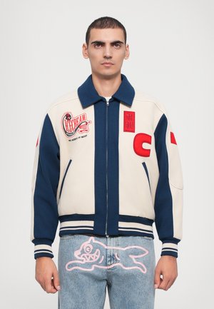 Creme- og marineblå varsity-jakke med lynlås, broderede patcher, ribbede manchetter og talje. Parret med blå jeans med pink grafiske detaljer.