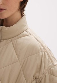 Osoba w beżowej pikowanej kurtce z widocznym przeszyciem i wysokim kołnierzem, przedstawiona od szyi do ramienia na białym tle.