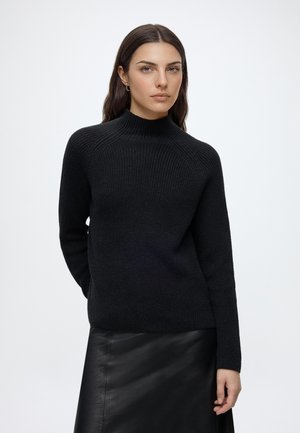 Vrouw met lang donker haar draagt een zwarte gebreide trui en een zwarte leren rok, staand tegen een eenvoudige lichte achtergrond.