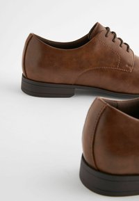 Chaussures en cuir marron avec une texture lisse, bout arrondi, design à lacets et une semelle en caoutchouc noir basse. Couture détaillée le long des bords.