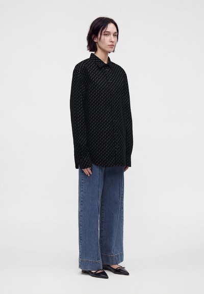Une personne se tient vêtue d'une chemise noire à manches longues à motifs, d'un jean bleu large et de ballerines noires pointues sur un fond blanc uni.