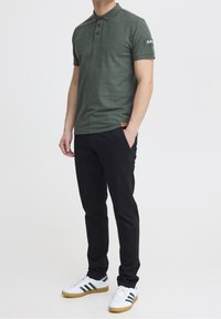 Zielona koszulka polo z trzema brązowymi guzikami, krótkimi rękawami i naszywką z logo. Połączona z czarnymi spodniami i białymi trampkami z zielonymi paskami.