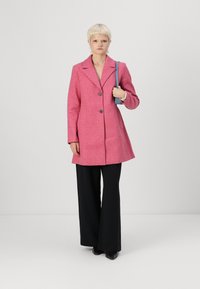 Roze wollen jas met een aangeknipte kraag, twee knopen en zijzakken, gedragen over zwarte wijde broek en gecombineerd met een blauwe schoudertas.