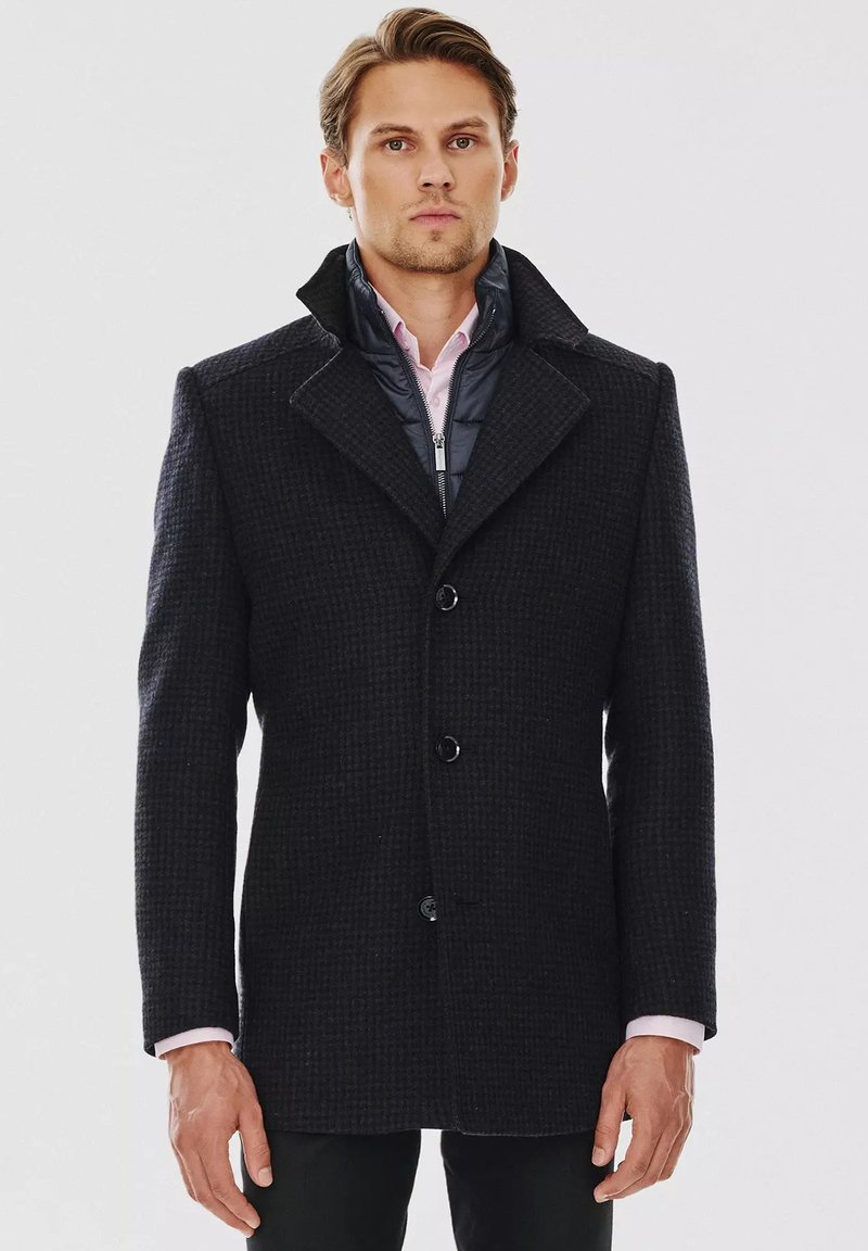 Czarny wełniany płaszcz o teksturowanym wzorze, z dużym kołnierzem, trzema guzikami, nałożony na granatową kurtkę puchową i jasnoróżową koszulę.