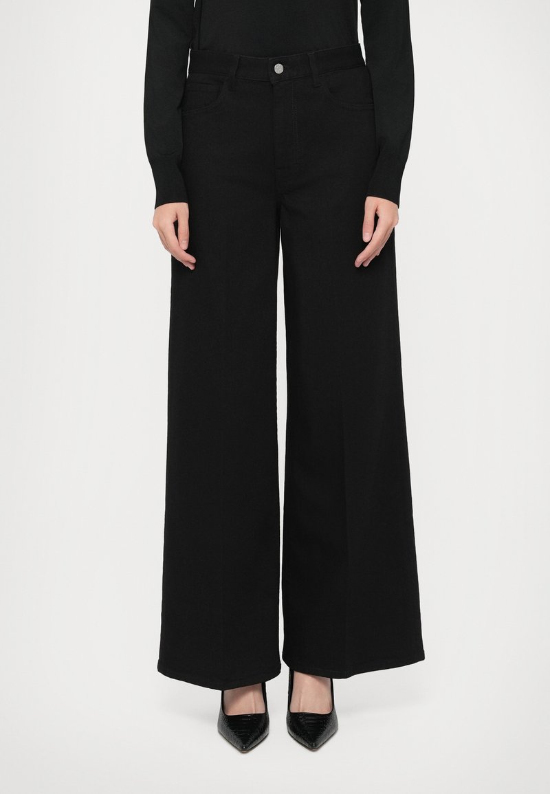 Pantaloni negri cu pantaloni evazați din țesătură, cu talie înaltă, având închidere cu buton frontal și buzunare, asortați cu pantofi negri cu vârf ascuțit.