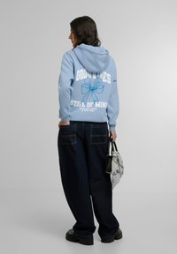 Lyseblå hettegenser med hvit tekst og blomstergrafikk på baksiden; kombinert med mørke denimbukser og bærende en svart og mønstrete veske.