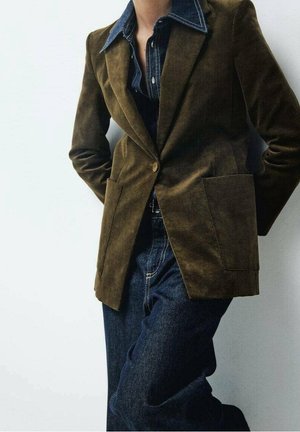 Henkilö, joka pukeutunut ruskeaan bleiseriin tumman farkkupaidan ja sinisten farkkujen päällä, seisoo kädet selän takana yksiväristä valkoista taustaa vasten.