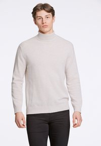 Maglione crema a collo alto in morbido tessuto texturizzato con maniche lunghe, scollo e orlo a costine. Indossato con pantaloni scuri aderenti.