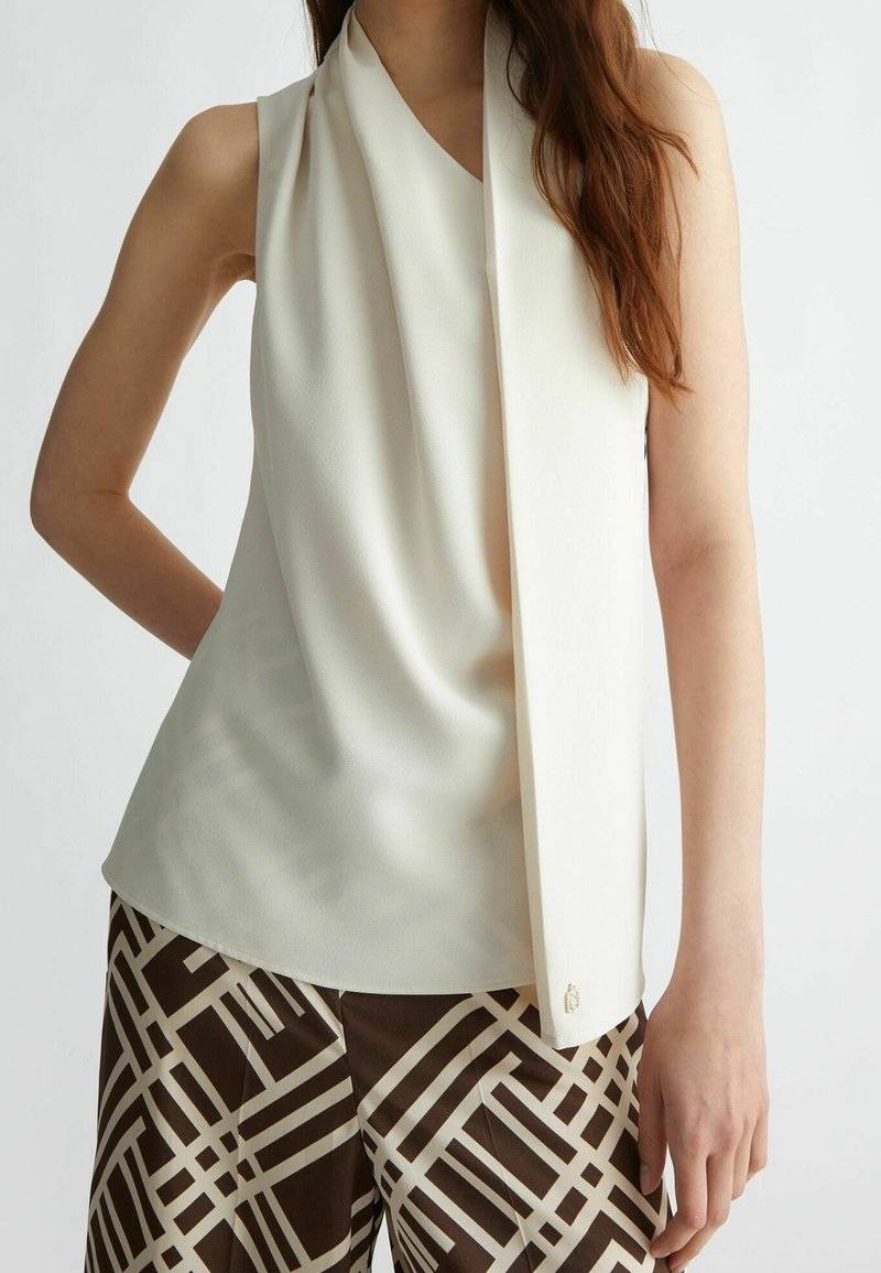 Top sans manches crème avec un design drapé et une texture douce. Associé à un pantalon marron à motifs présentant des lignes géométriques et des accents.