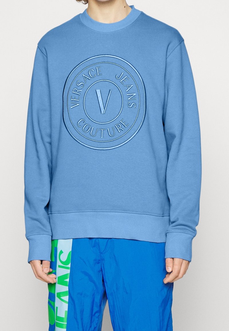 Personne portant un sweat-shirt bleu clair avec le logo "Versace Jeans Couture" et un pantalon bleu vif avec des panneaux latéraux verts et blancs.