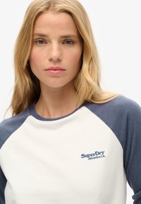 Witte en marineblauwe raglan sweatshirt met het geborduurde "SuperDry" logo op de borst. Zachte stof met een ronde hals.