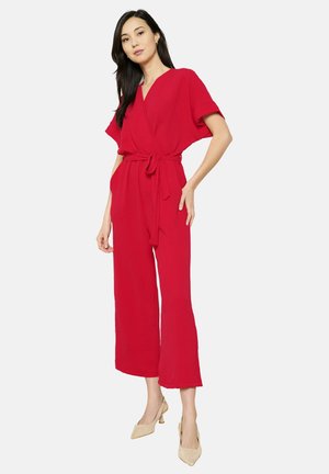 Roter Jumpsuit mit kurzen Ärmeln, tailliertem Gürtel, weitem Bein und strukturiertem Stoff. Kombiniert mit beigen High Heels.