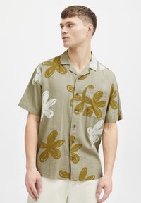 Camisa de manga corta en verde oliva, con grandes patrones florales en blanco y marrón, cierre de botones en la parte delantera y un bolsillo en el pecho.