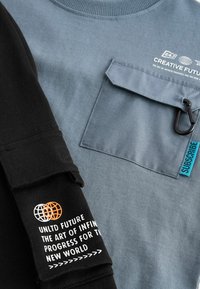 Zweifarbiger Sweatshirt mit grauem Körper und schwarzen Ärmeln. Verfügt über eine Brusttasche, Textgrafiken und ein blaues Etikett mit "ABONNIEREN."