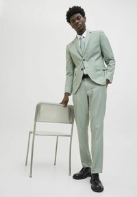 Abito verde chiaro con un design su misura, caratterizzato da una giacca a un bottone e pantaloni slim. Abbinato a una camicia bianca e una cravatta chiara. Scarpe nere.