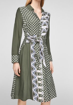 Kaulusmekko oliivinvihreänä, jossa pilkkuja, vyötäröllä solmittava kietaisunauha ja sinivalkoiset kuvioidut paneeliyksityiskohdat. Pitkät hihat.