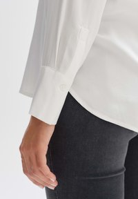 Camicia bianca a maniche lunghe con polsini abbottonati, che mostra una trama liscia. Abbinate a jeans scuri, che evidenziano un taglio classico e su misura.